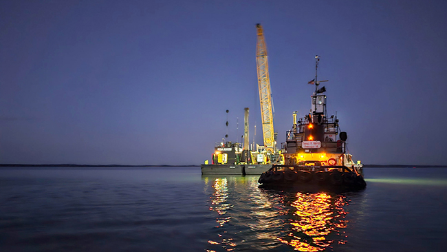 A nighttime photo of a tug and barge on calm water. Both are lit with work lights and are reflecting on the water, which has slight ripples.